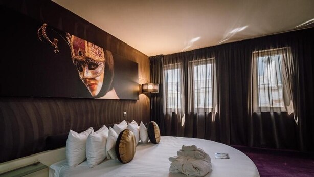 Une chambre d’hôtel avec un grand lit, une tête de lit ornée d’un élément décoratif, une œuvre d’art accrochée au mur et une fenêtre avec des rideaux.