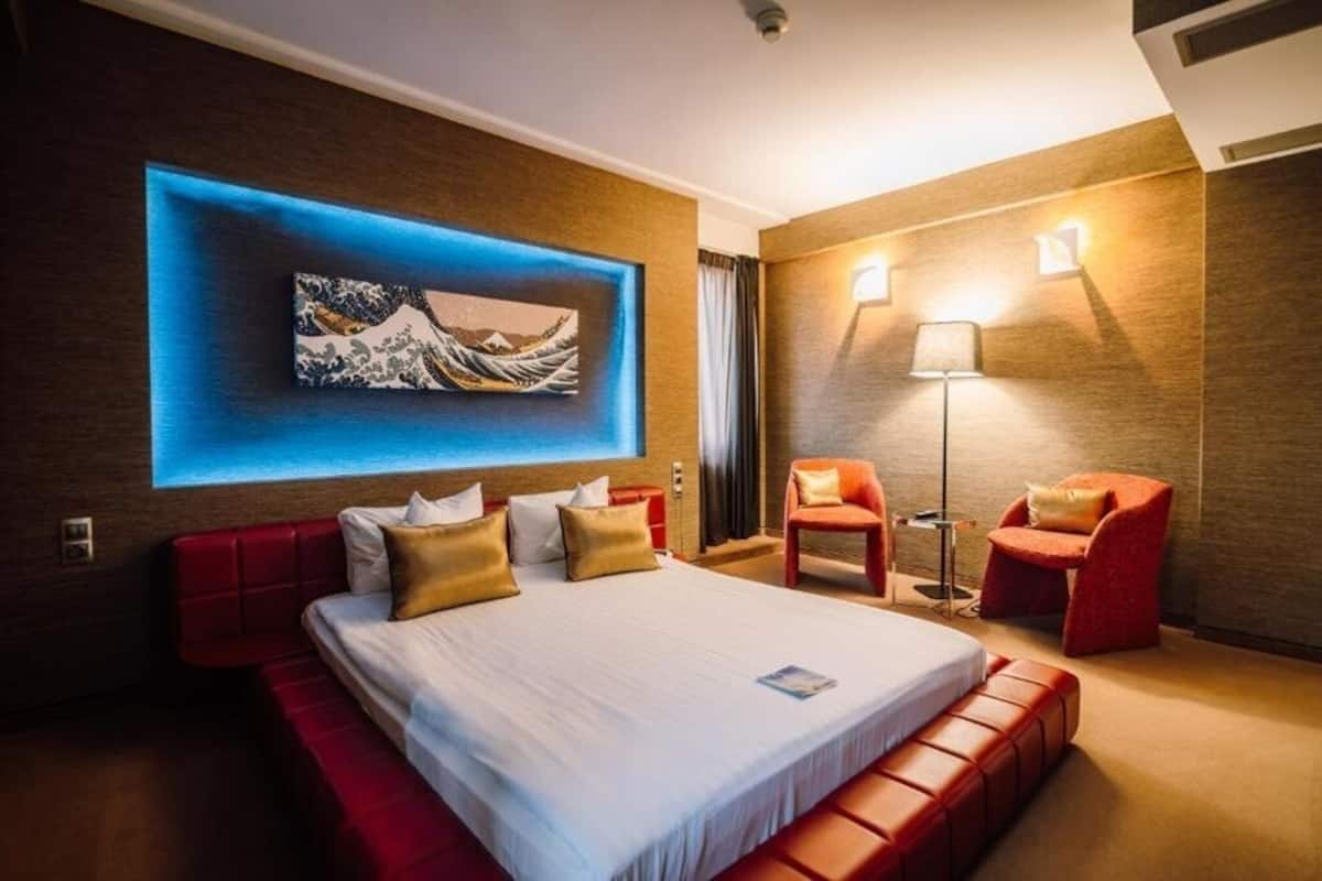 A modern hotel room with a large bed, red seating, a wall-mounted artwork, and a floor lamp.