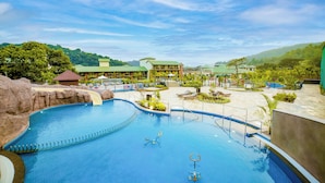 A swimming pool with a water slide, lounge chairs, and a building with a green roof in the background. - Club Mahindra Assonora (Assonora)