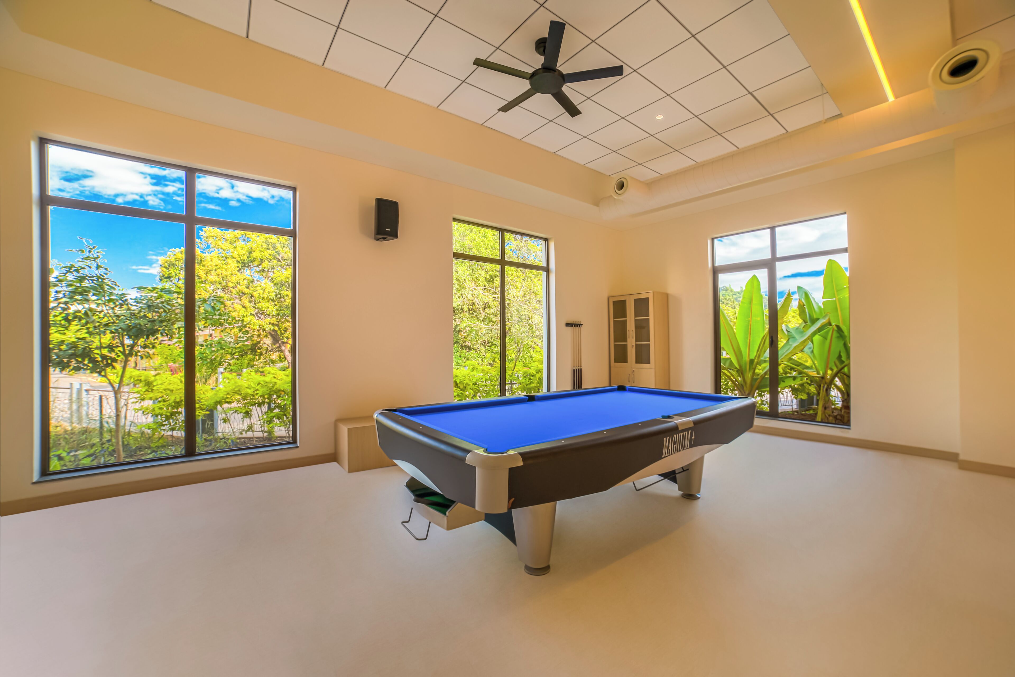 Une table de billard dans une pièce dotée de grandes fenêtres, d’un ventilateur de plafond et offrant une vue sur la verdure.