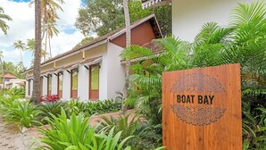 Un letrero de madera con la inscripción 'Boat Bay', rodeado de exuberante follaje y un edificio tradicional con techo de tejas.