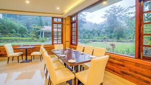 A dining area with a long table set for six, surrounded by white chairs, large windows with wooden frames, and a view of a lush garden. - Club Mahindra Le Vintuna (Gangtok)