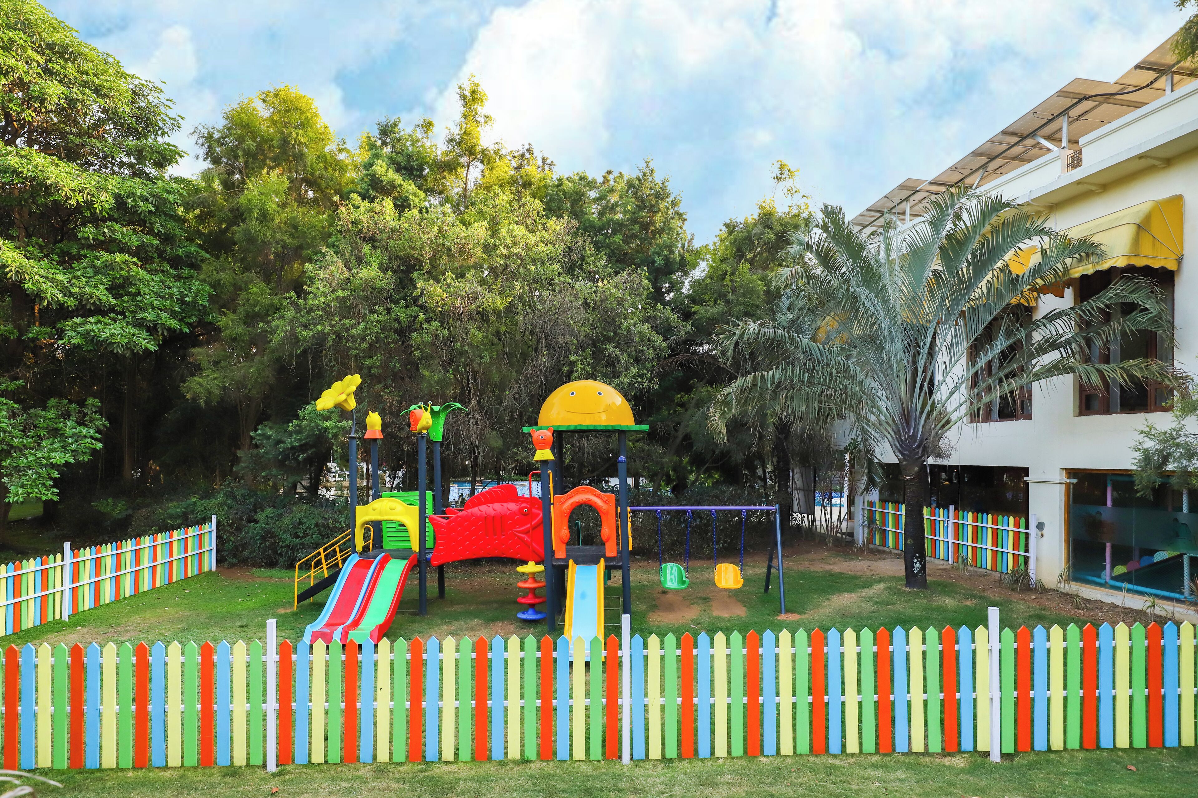 A playground with slides and climbing structures, surrounded by a white picket fence and lush greenery.