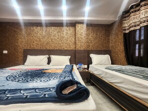 A hotel room with two beds, a patterned bedspread, a rolled-up blanket, a nightstand with a blue container, and a wall with a textured pattern. - Hotel The Divine (New Delhi)