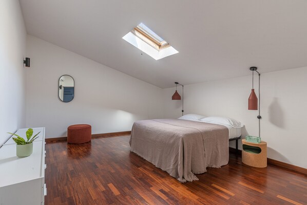 Une chambre avec un plafond en pente, un puits de lumière, un lit, une table de chevet, un miroir, une lampe rouge et une petite plante.