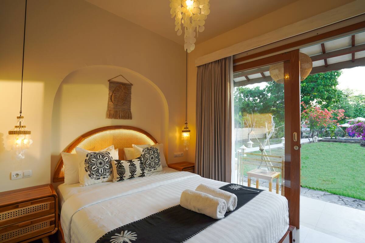 A bedroom with a large bed, a wooden headboard, a nightstand, a wall-mounted lamp, and a view of a garden through a sliding glass door.