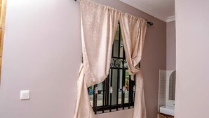 Une pièce avec une fenêtre recouverte de rideaux beiges, un mur blanc et une salle de bain équipée d’un lavabo et d’un miroir.