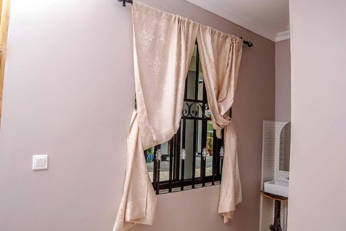 A room with a window covered by beige curtains, a white wall, and a bathroom with a sink and mirror.