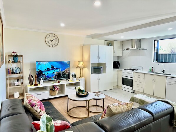 A modern living room with a flat-screen TV, sofa, coffee table, and a kitchen area with cabinets, microwave, and stove.