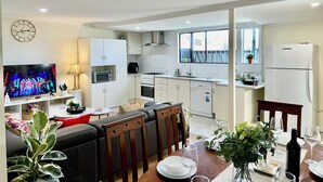 A modern kitchen with white cabinets, a stove, microwave, and dishwasher. There is a dining area with a table set for two, a sofa, and a TV in the background. - Woodville Garden Home in great location (Woodville Gardens)