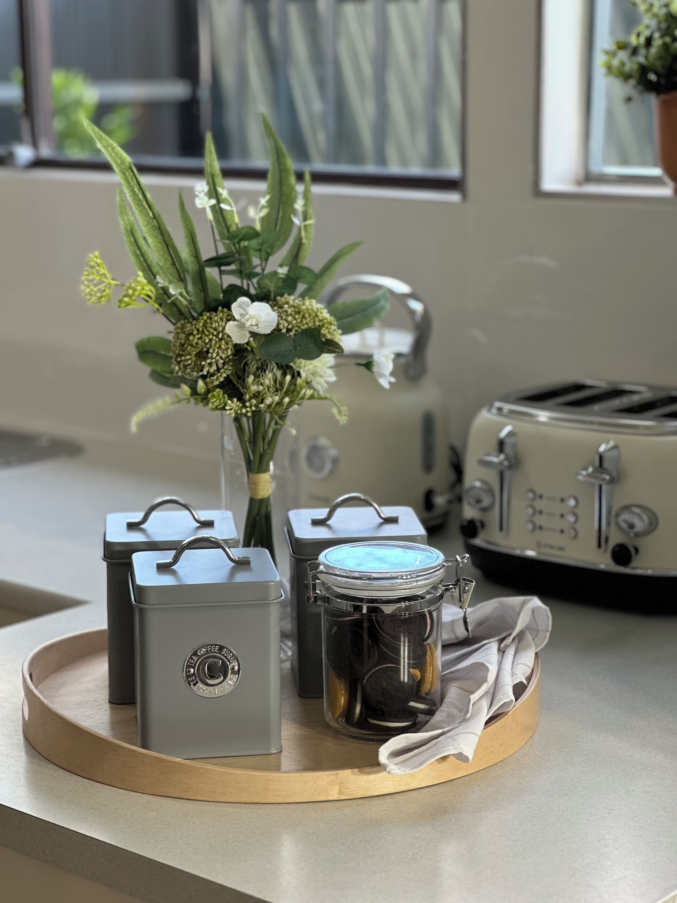 Un comptoir de cuisine sur lequel se trouve un plateau avec une théière, un bocal de biscuits et un vase rempli de fleurs.
