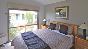 A bedroom with a large bed, a window, a bedside table with a lamp, and a painting on the wall. - 12 Raheen Drive, Mallacoota (Mallacoota)