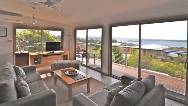 A modern living room with a large sofa, a coffee table, and a TV on a wooden stand. There is a balcony with a view of the sea and greenery. - 12 Raheen Drive, Mallacoota (Mallacoota)