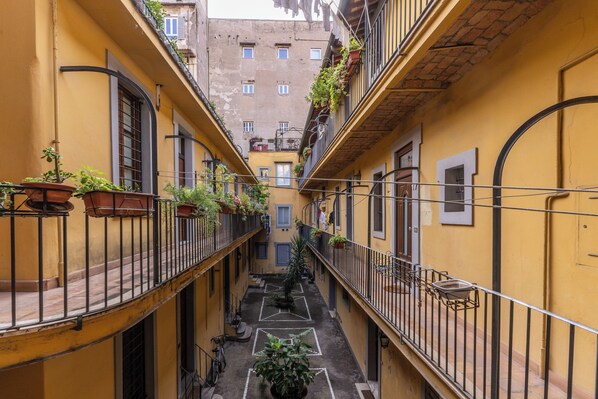 A narrow alleyway with yellow buildings, balconies with potted plants, and a central pathway marked with geometric shapes.