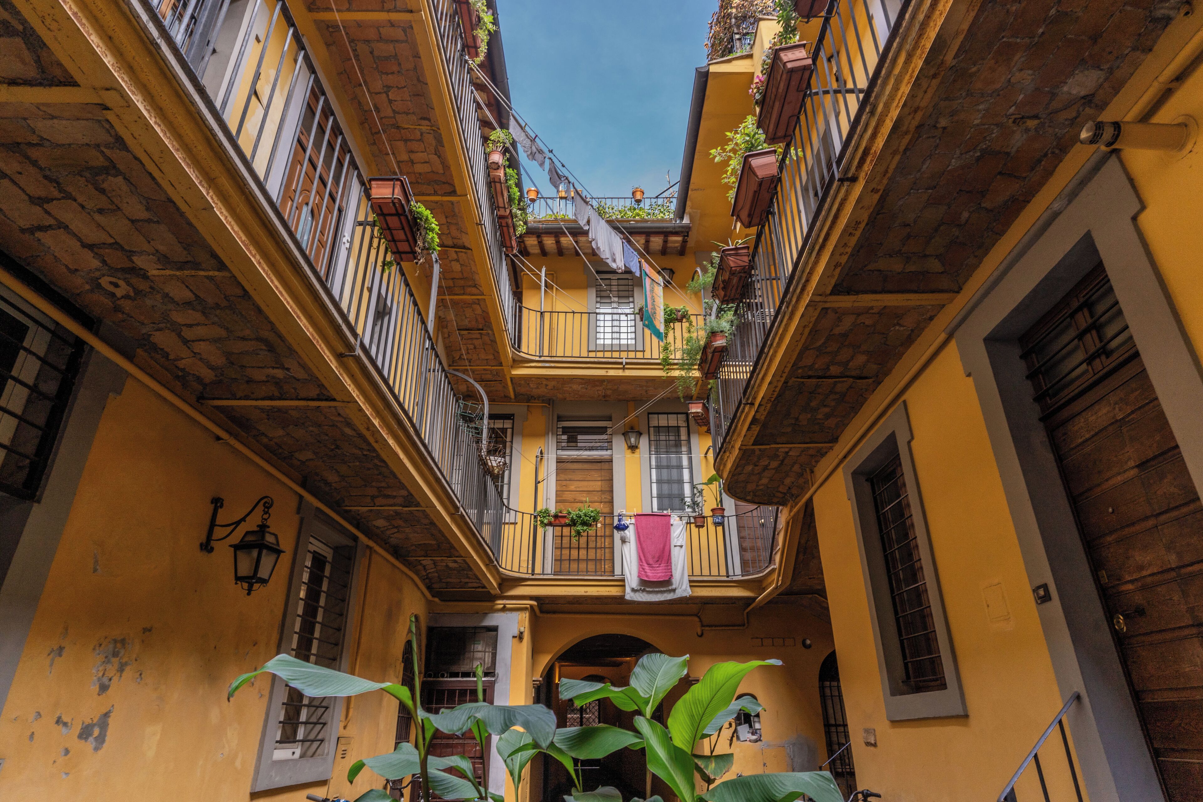 A narrow alleyway with yellow buildings, balconies with plants, and a hanging laundry line.