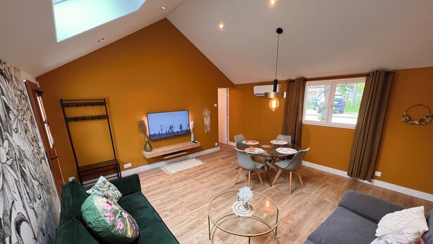 A modern living room with a high ceiling, a flat-screen TV mounted on the wall, a dining area with a table and chairs, and a sofa with floral cushions.