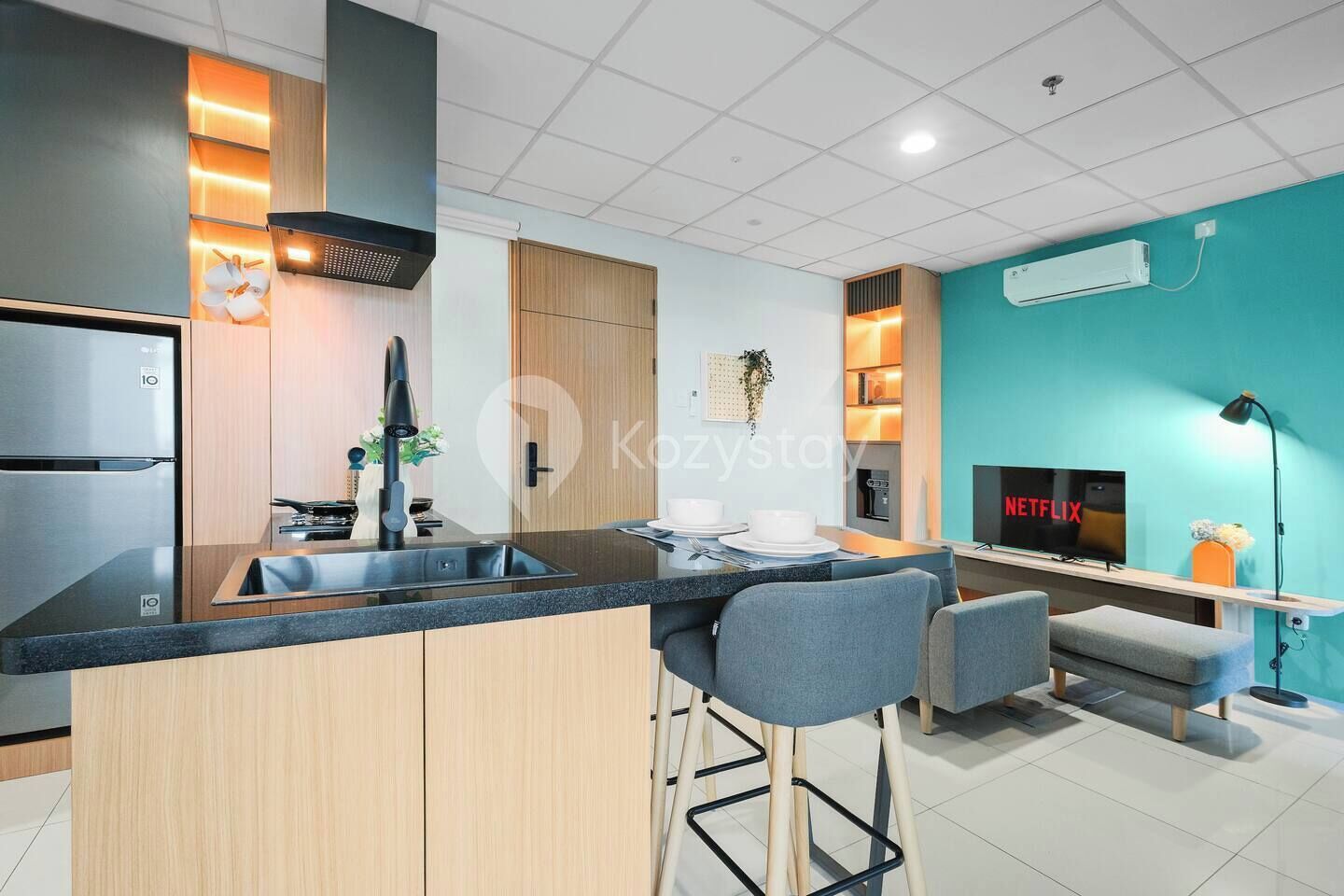 A modern kitchen with a central island, a built-in stove, and a sink. There is a dining area with a TV and a sofa.