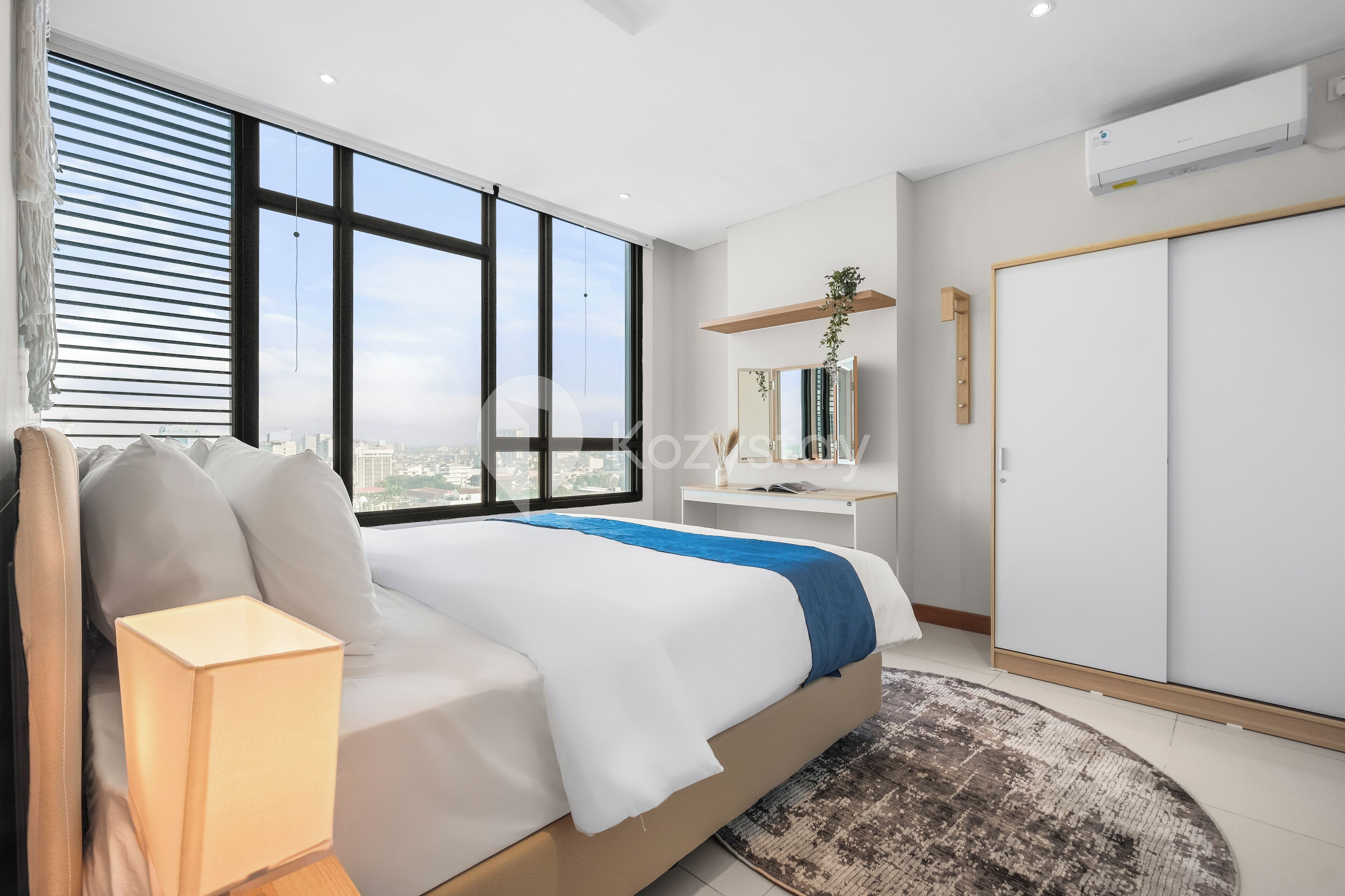 Un dormitorio moderno con una cama grande, una lámpara de noche, vistas de la ciudad a través de ventanales del suelo al techo, un escritorio pequeño y un armario.
