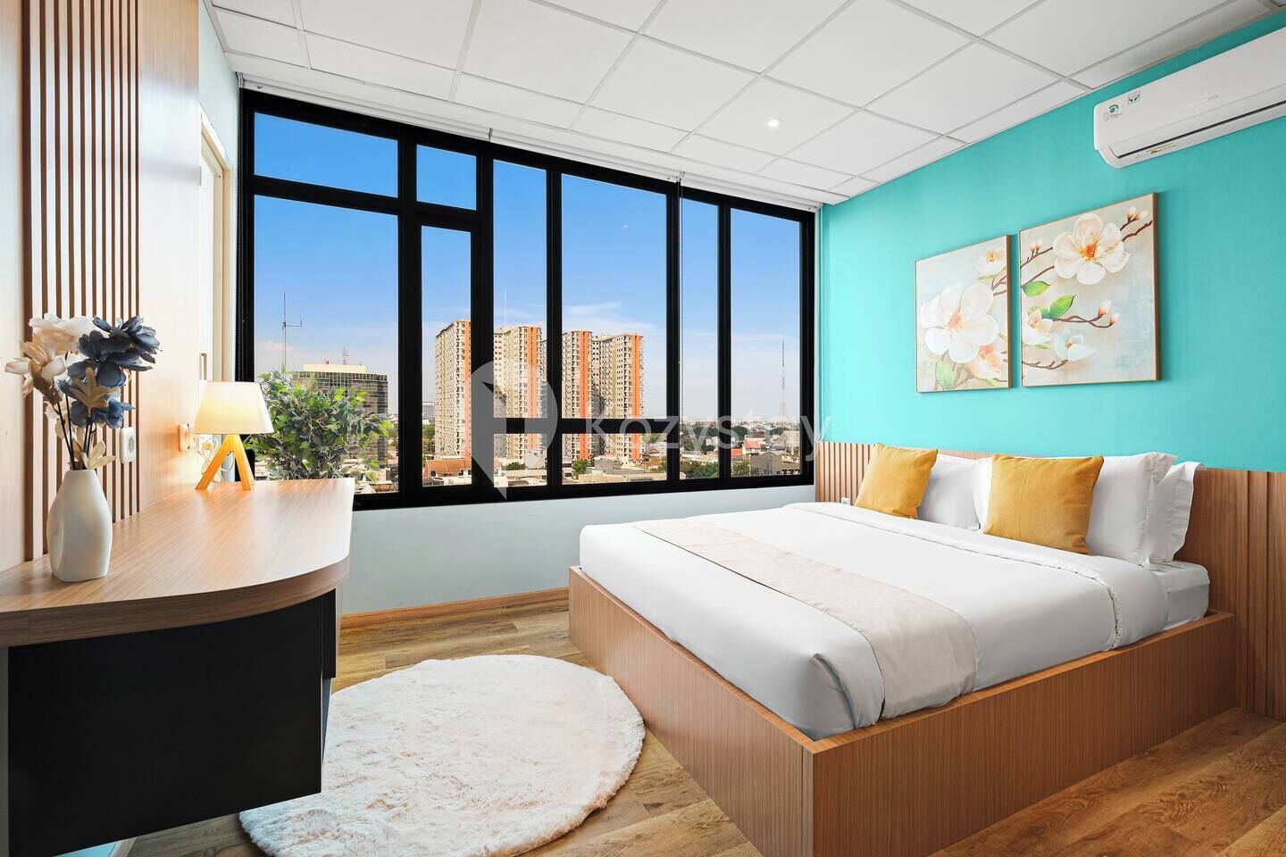 A modern bedroom with a large window, a bed with white bedding, a wooden bedside table, and a small rug.