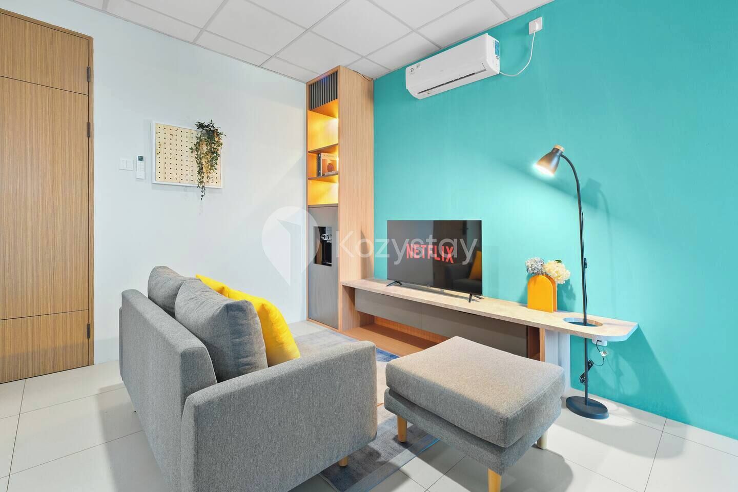 A modern living room with a grey sofa, a small ottoman, a TV on a stand, and a wall-mounted air conditioner.