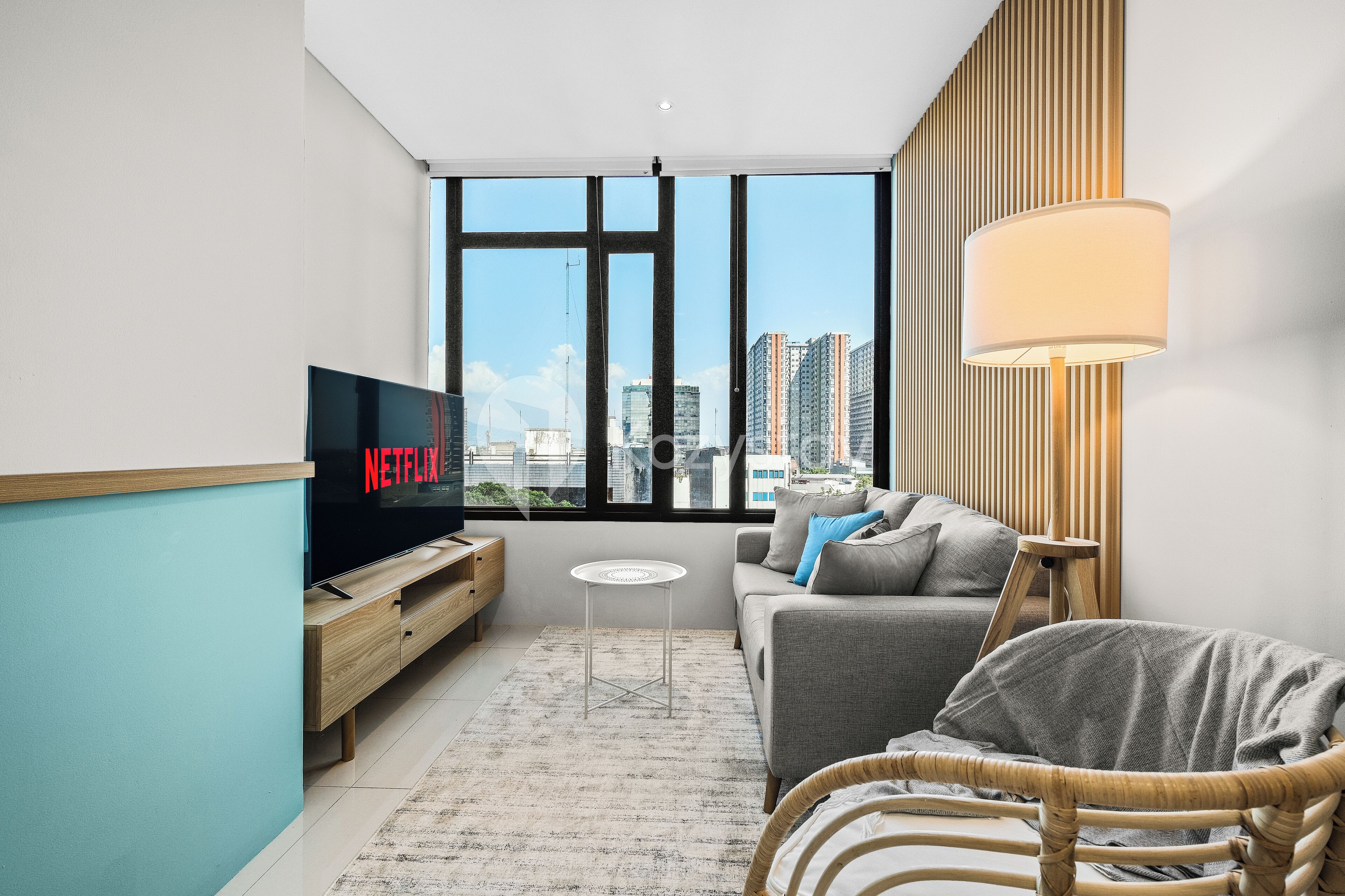 Ein modernes Wohnzimmer mit einem Fernseher, der 'NETFLIX' anzeigt, einem Sofa, einem kleinen Tisch und Blick auf die Stadt durch große Fenster.