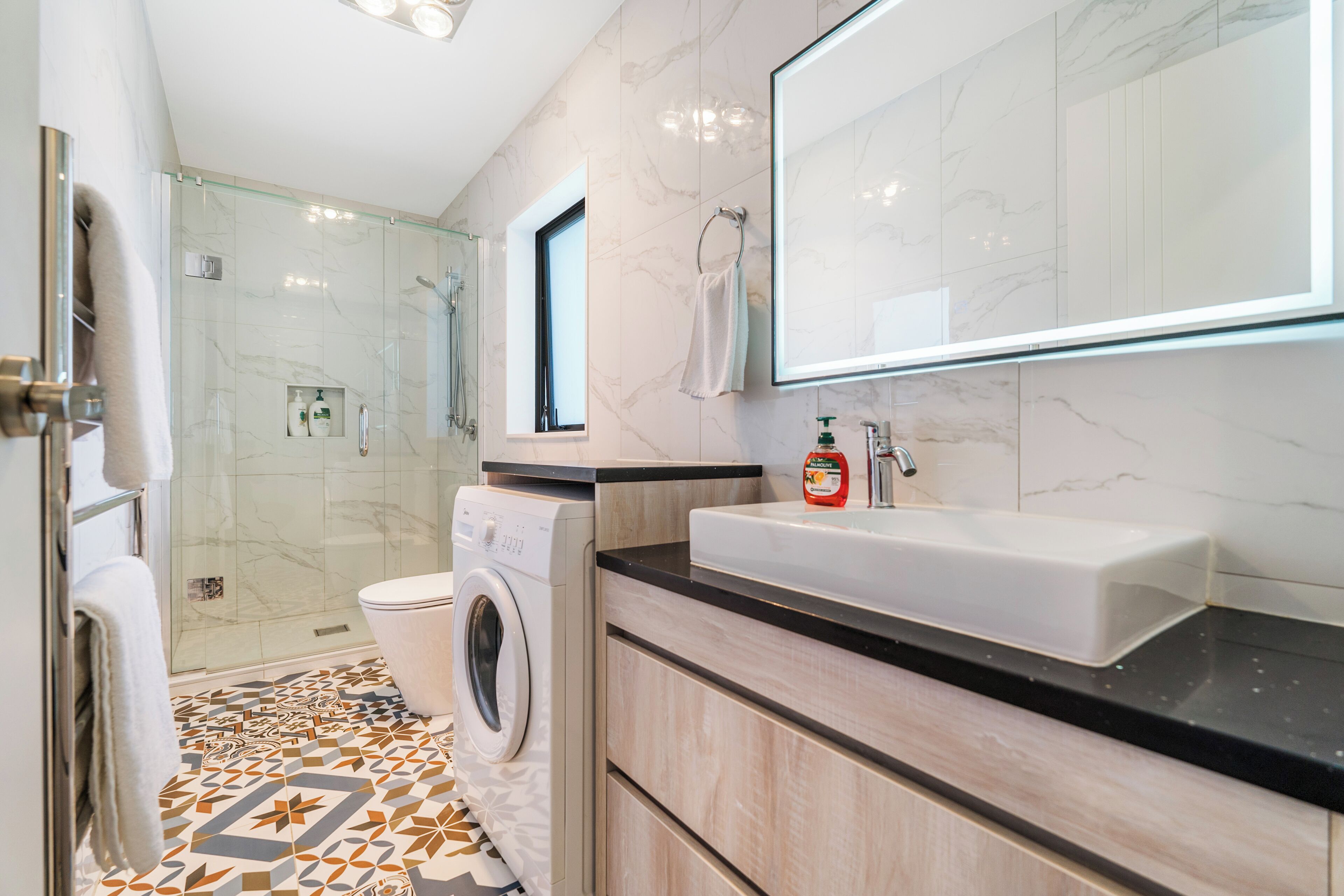 A modern bathroom with a glass-enclosed shower, a built-in washing machine, a sink with a soap dispenser, and a large mirror.