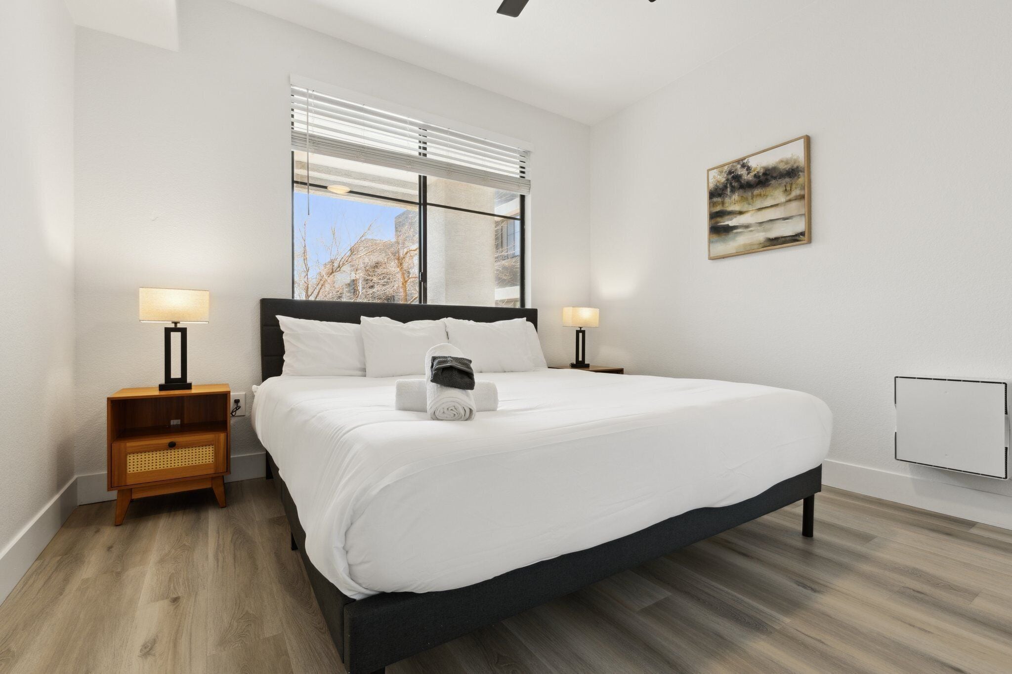 Una camera da letto moderna con un letto grande, due comodini con lampade, una finestra con persiane e un quadro appeso al muro.