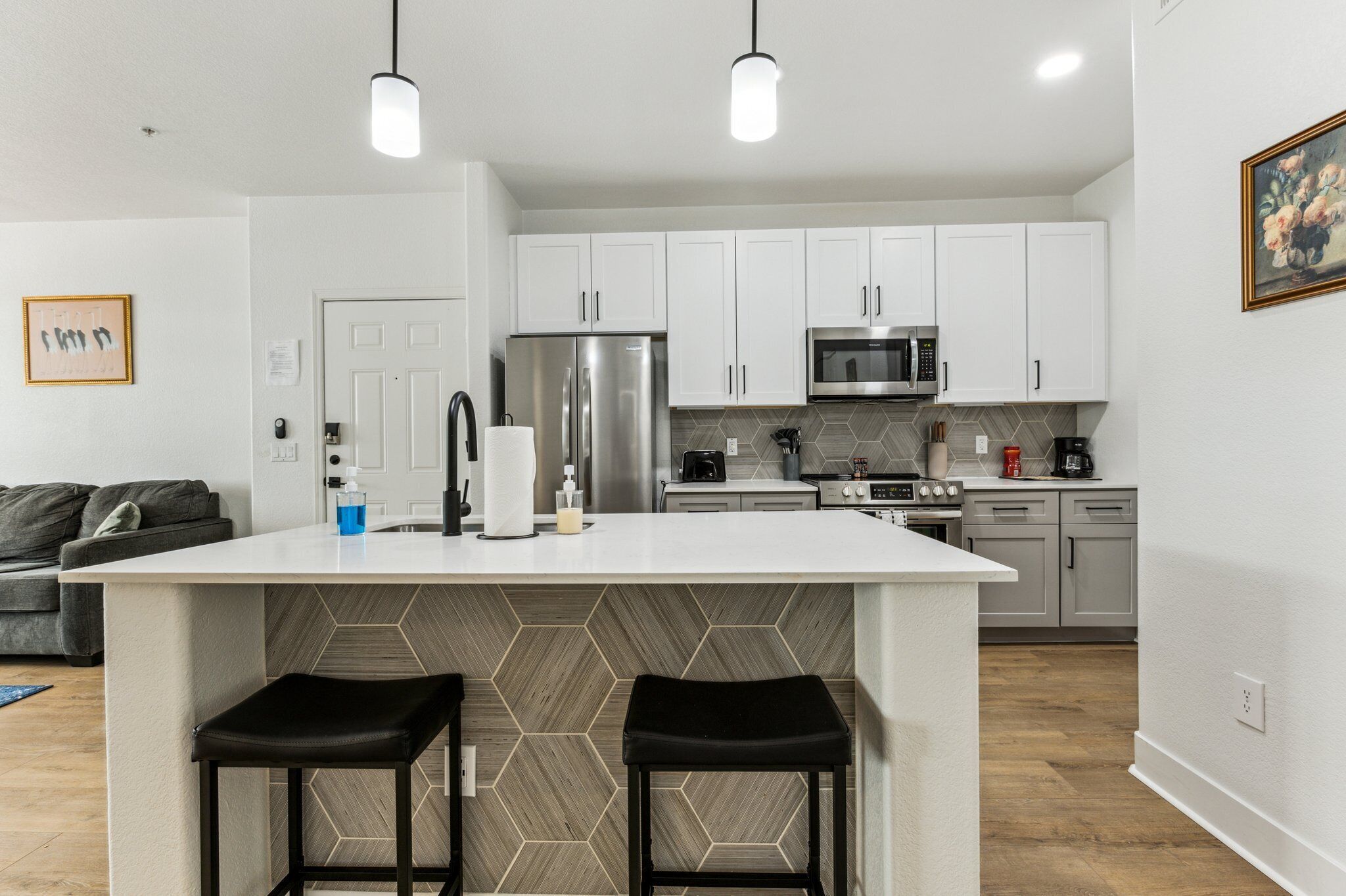 Una cocina moderna con armarios blancos, una isla central con taburetes, electrodomésticos de acero inoxidable y un salpicadero de azulejos tipo espiga.