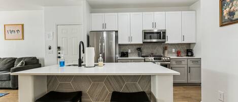 Une cuisine moderne avec des meubles blancs, un îlot central doté de tabourets, des appareils en acier inoxydable et un dosseret de carrelage disposé en motif chevrons.