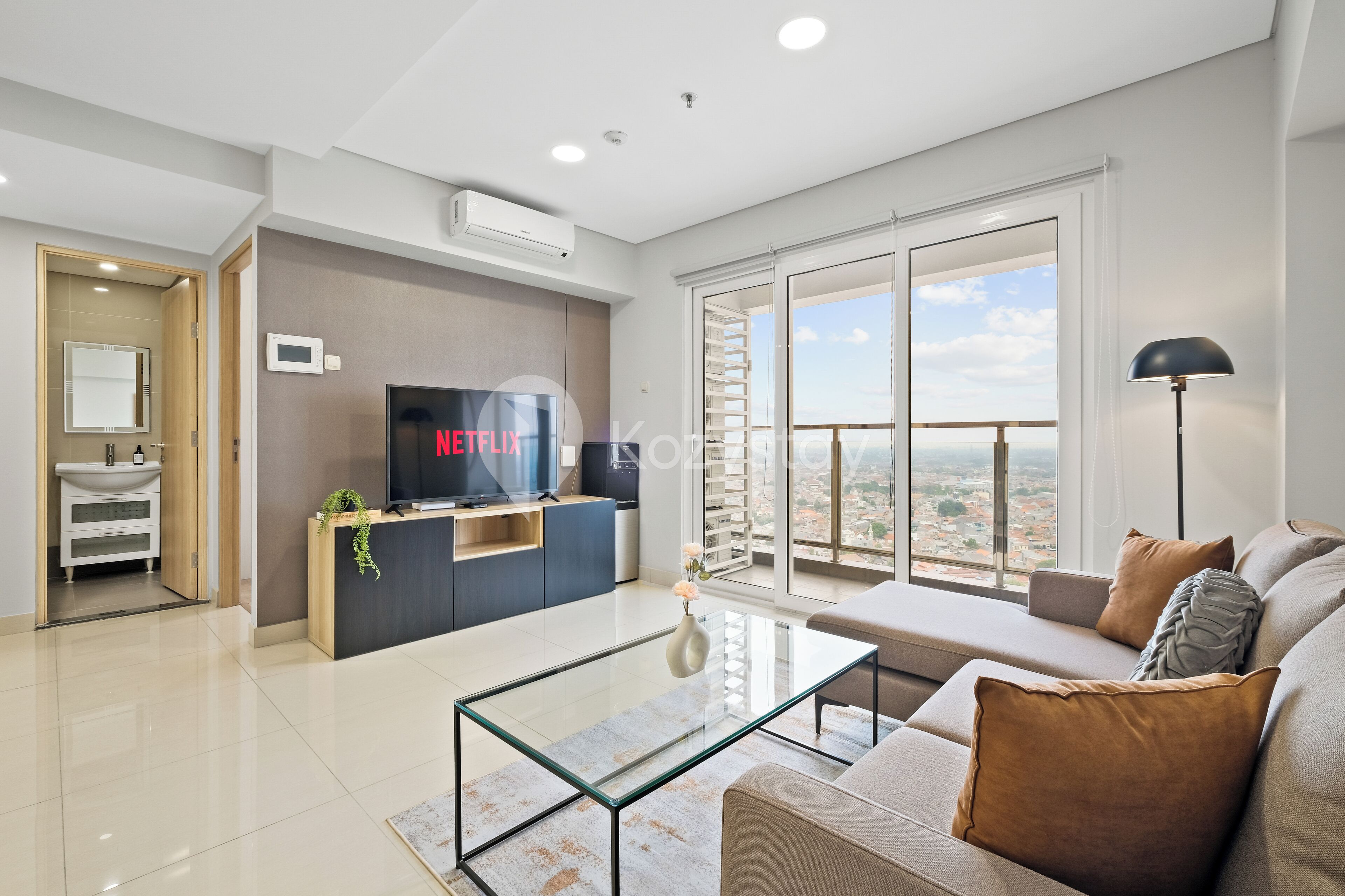 Un moderno salón con una mesa de centro de vidrio, un sofá con cojines, un televisor y un balcón con vistas a la ciudad.
