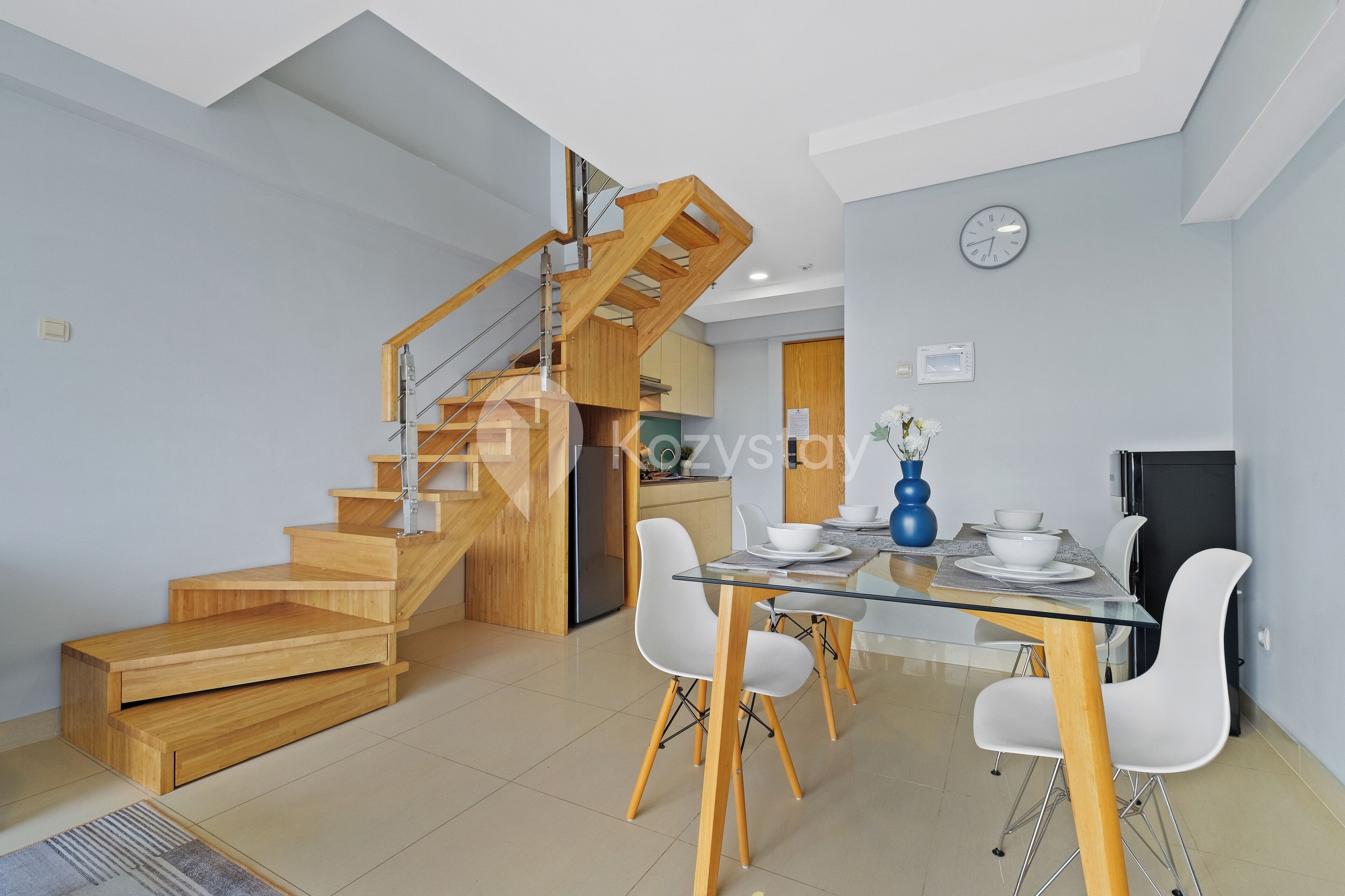 Uma cozinha moderna com escada de madeira, área de jantar com mesa de vidro e cadeiras brancas.