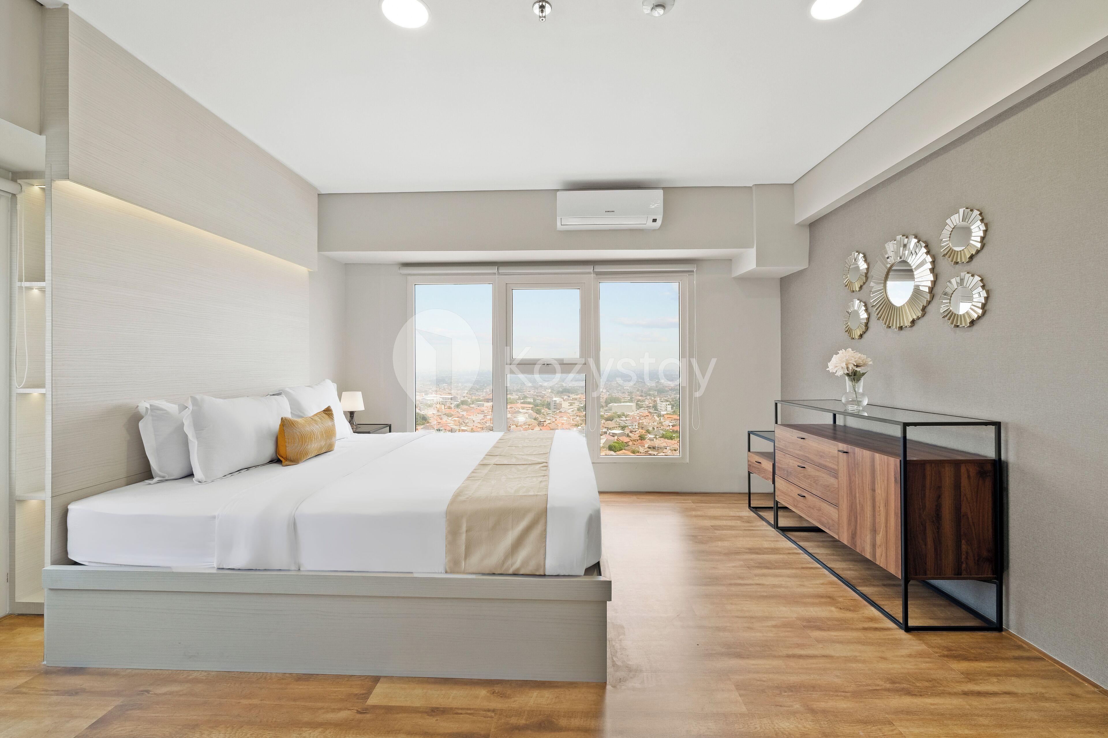 Ein modernes Schlafzimmer mit einem großen Bett, einer hölzernen Kommode und Blick auf die Stadt durch große Fenster.