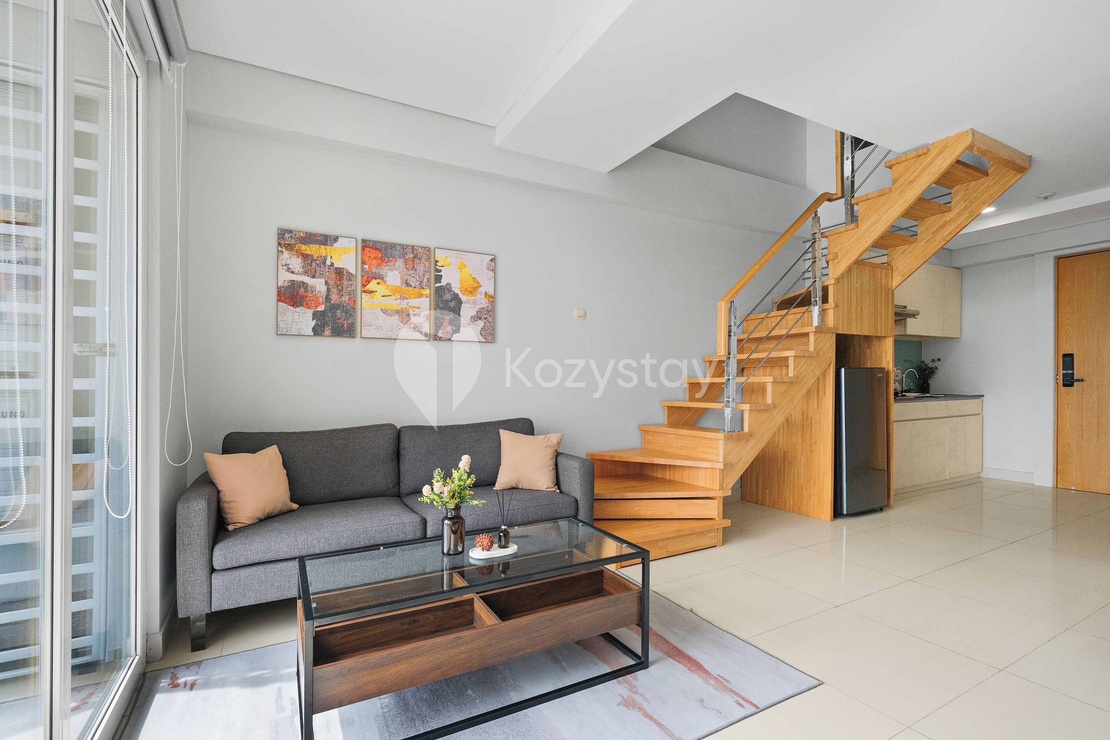 A modern living room with a grey sofa, a glass coffee table, and a wooden staircase.