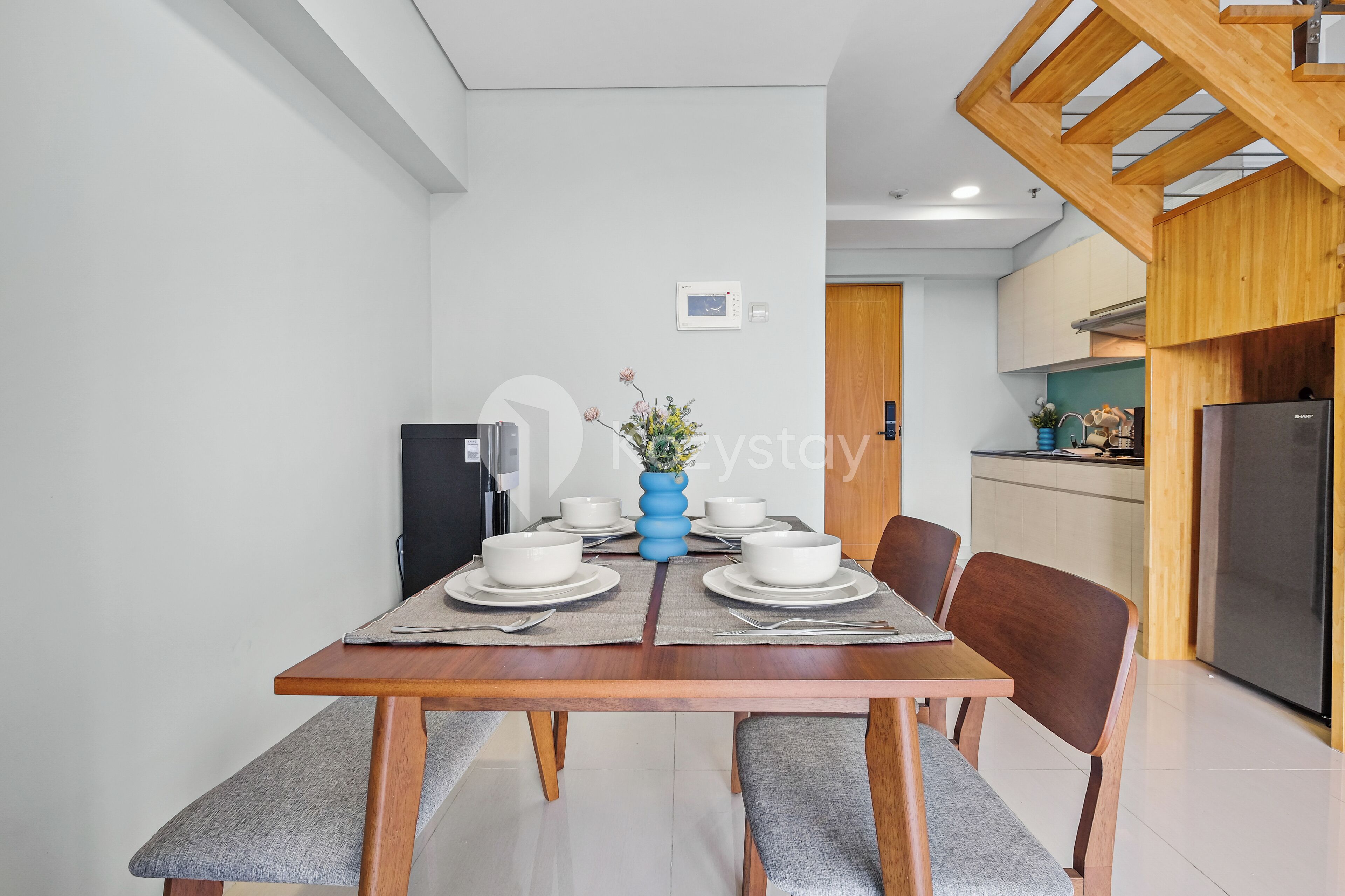 A modern dining area with a wooden table set for four, a blue vase centerpiece, and a kitchen in the background.