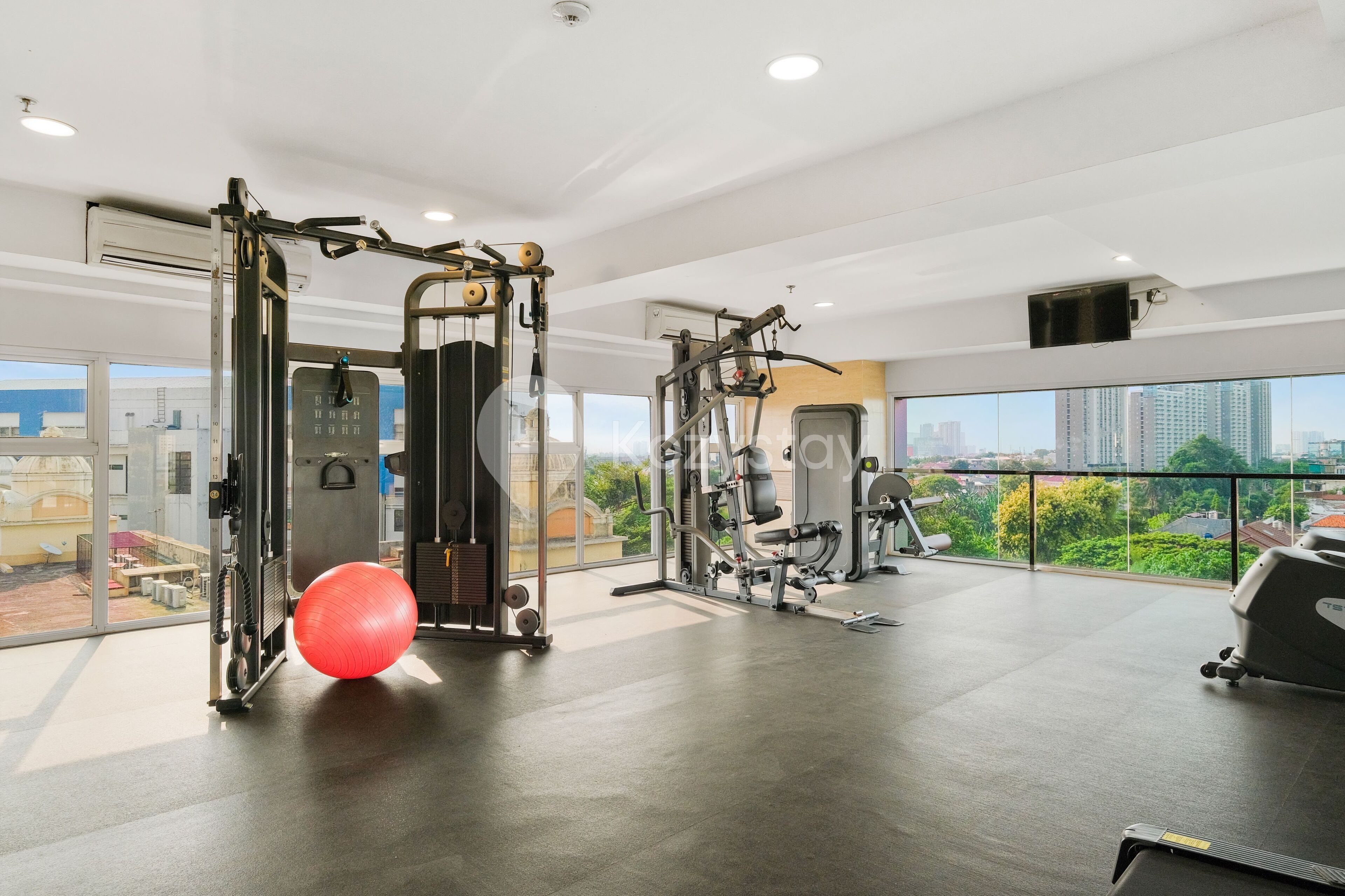 A modern gym with various exercise equipment, large windows offering city views, and a red exercise ball.