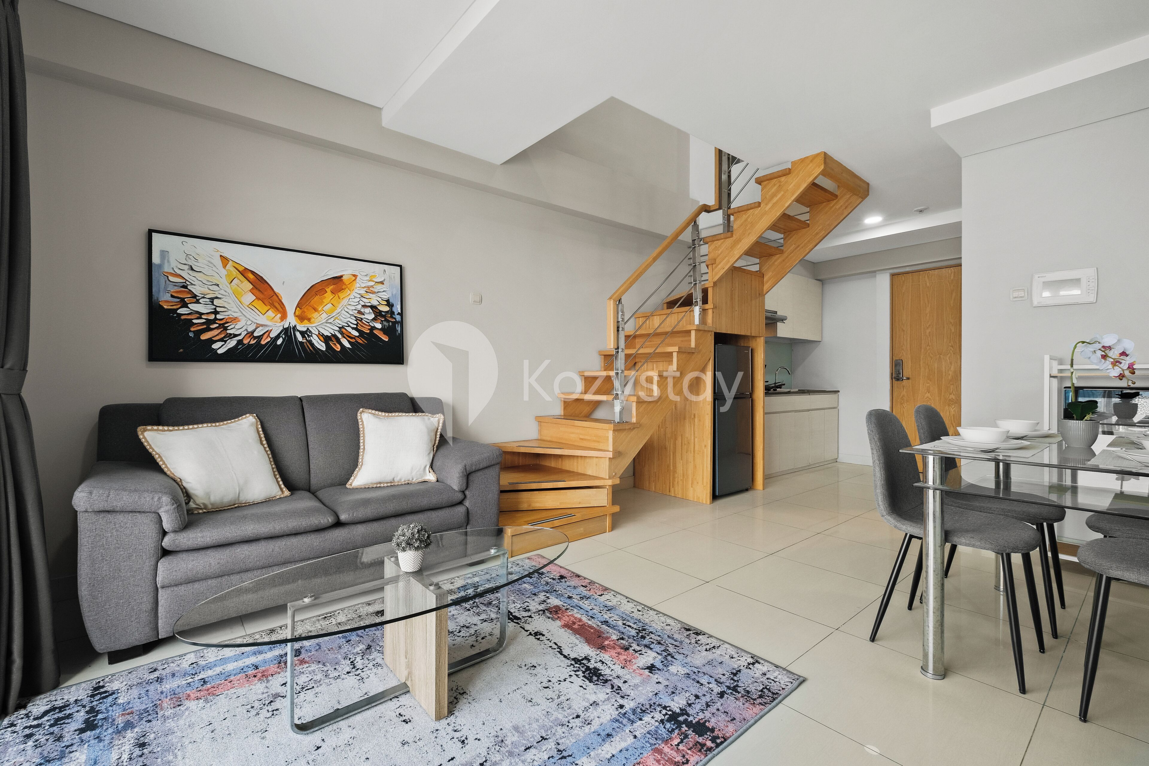 A modern living room with a grey sofa, a glass coffee table, and a staircase leading to a kitchen.