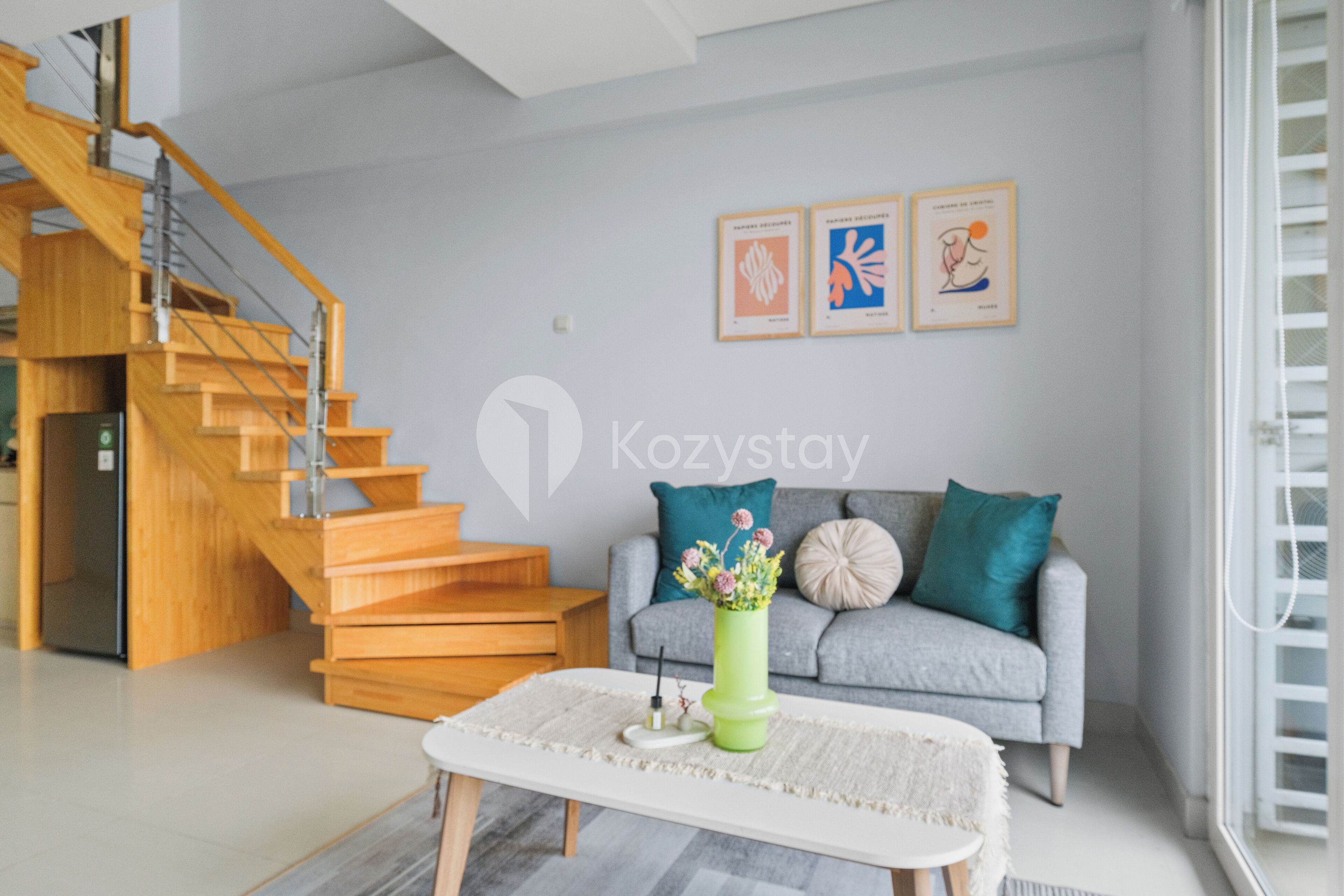 A modern living room with a staircase, grey sofa, and artwork on the wall.