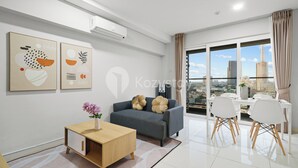 A modern living room with a grey sofa, wooden coffee table, and white chairs. There are abstract paintings on the wall and a large window with a city view. - Magnolia Aparthotel by Kozystay - Kebon Jeruk (Jakarta)
