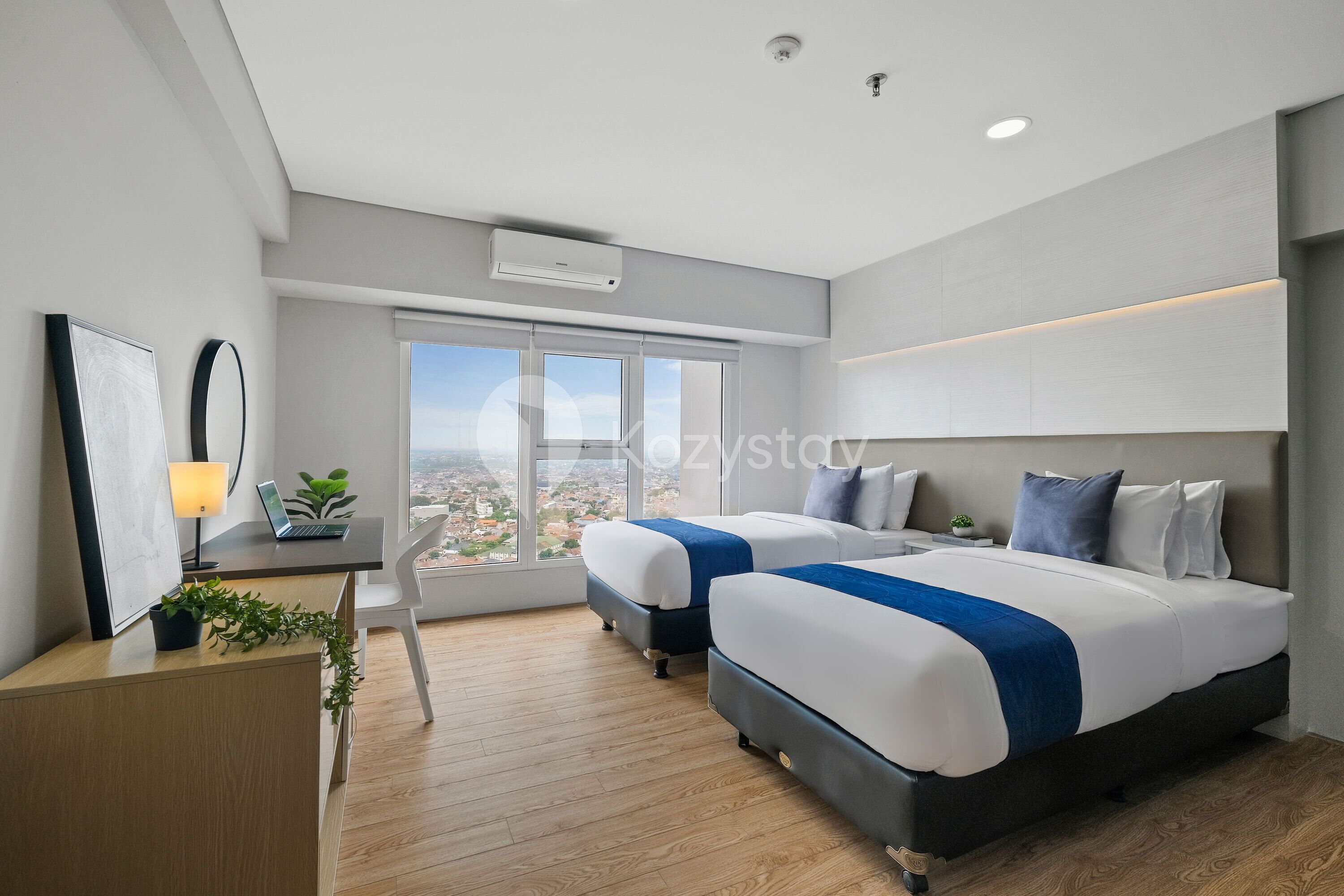 A modern hotel room with two beds, a desk with a laptop, a potted plant, and a large window offering a city view.