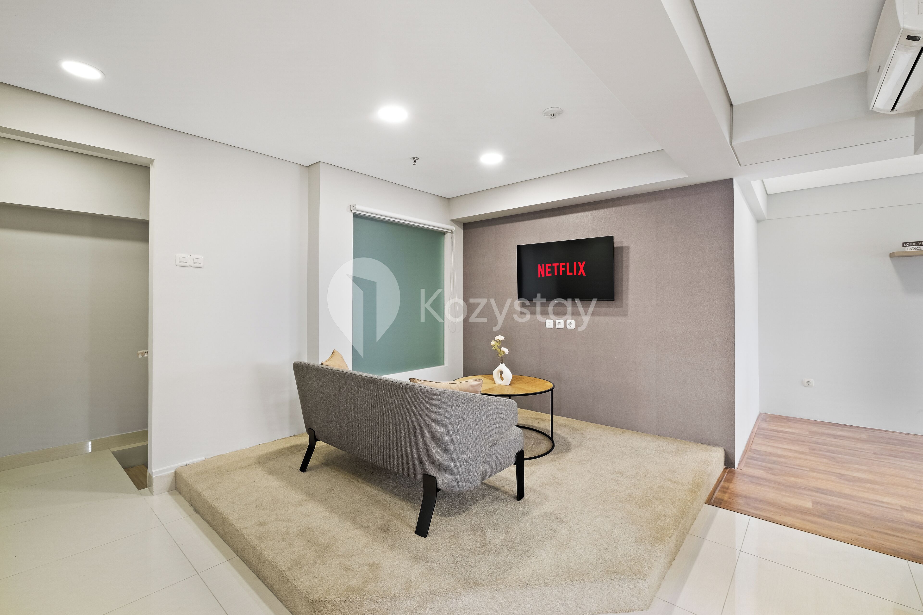A modern, well-lit room with a grey sofa, a small round table, and a wall-mounted TV displaying 'NETFLIX'.