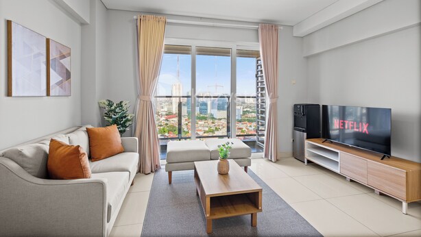 A modern living room with a sofa, a coffee table, a TV, and a view of the cityscape through large windows. - Magnolia Aparthotel by Kozystay - Kebon Jeruk (Jakarta)