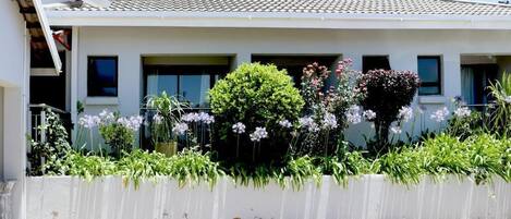 Una casa de dos pisos con techo de tejas, fachada blanca y un jardín con diversas plantas y flores.