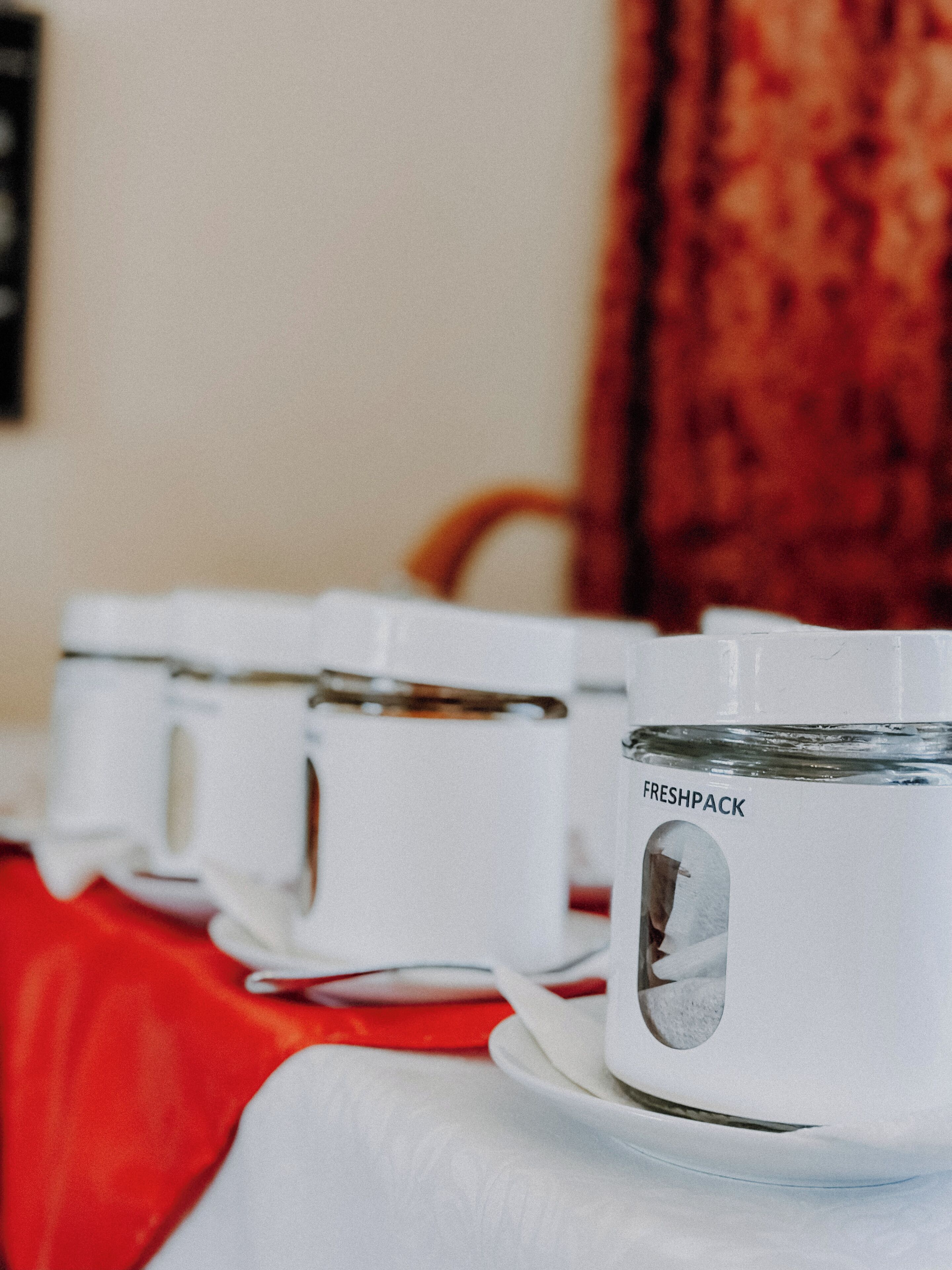 Una hilera de tazas blancas con la etiqueta 'FRESHPACK' sobre una mesa con mantel rojo.