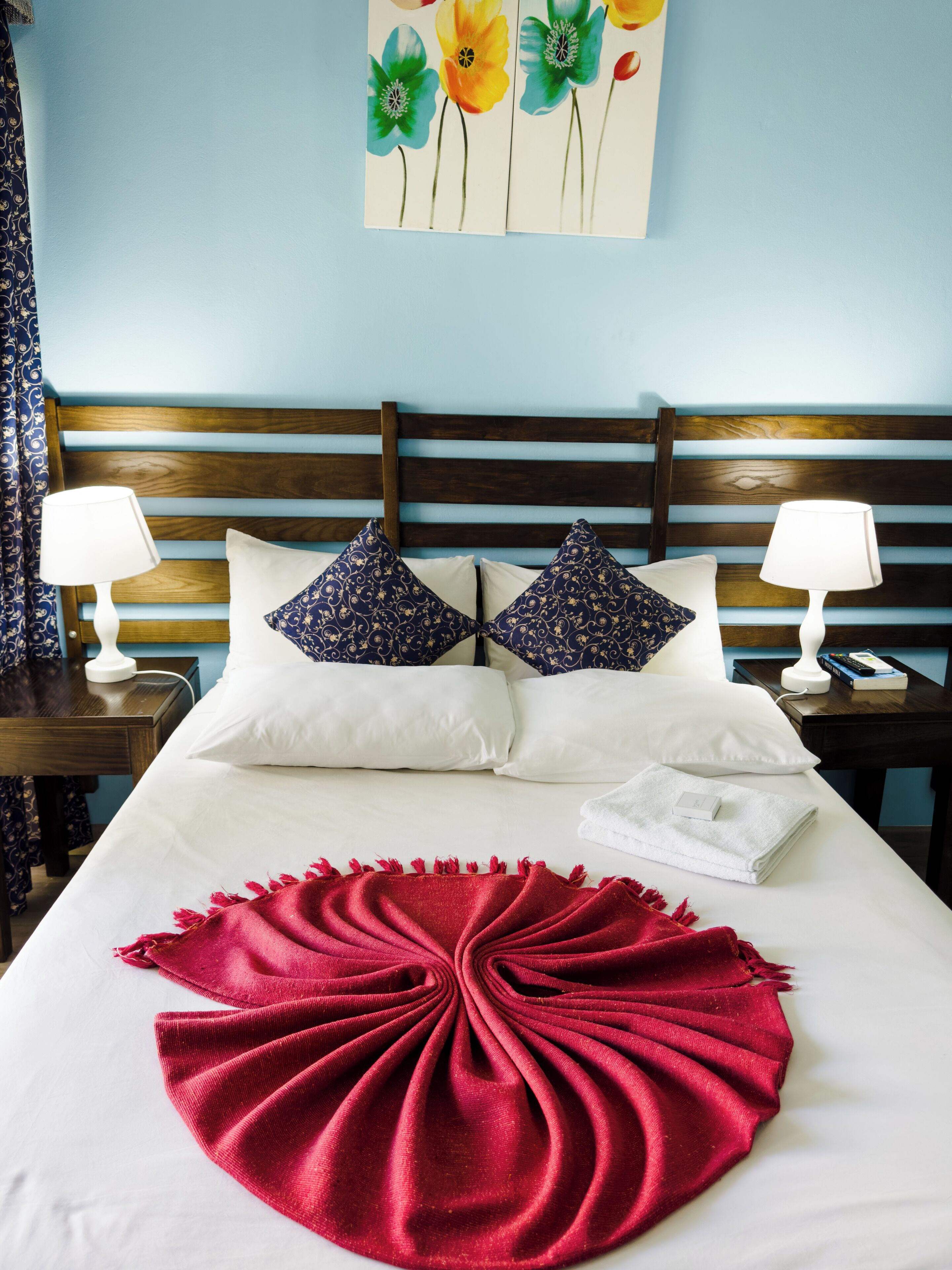 A neatly made bed with white and blue patterned pillows, a red throw blanket, and a wall art of flowers.