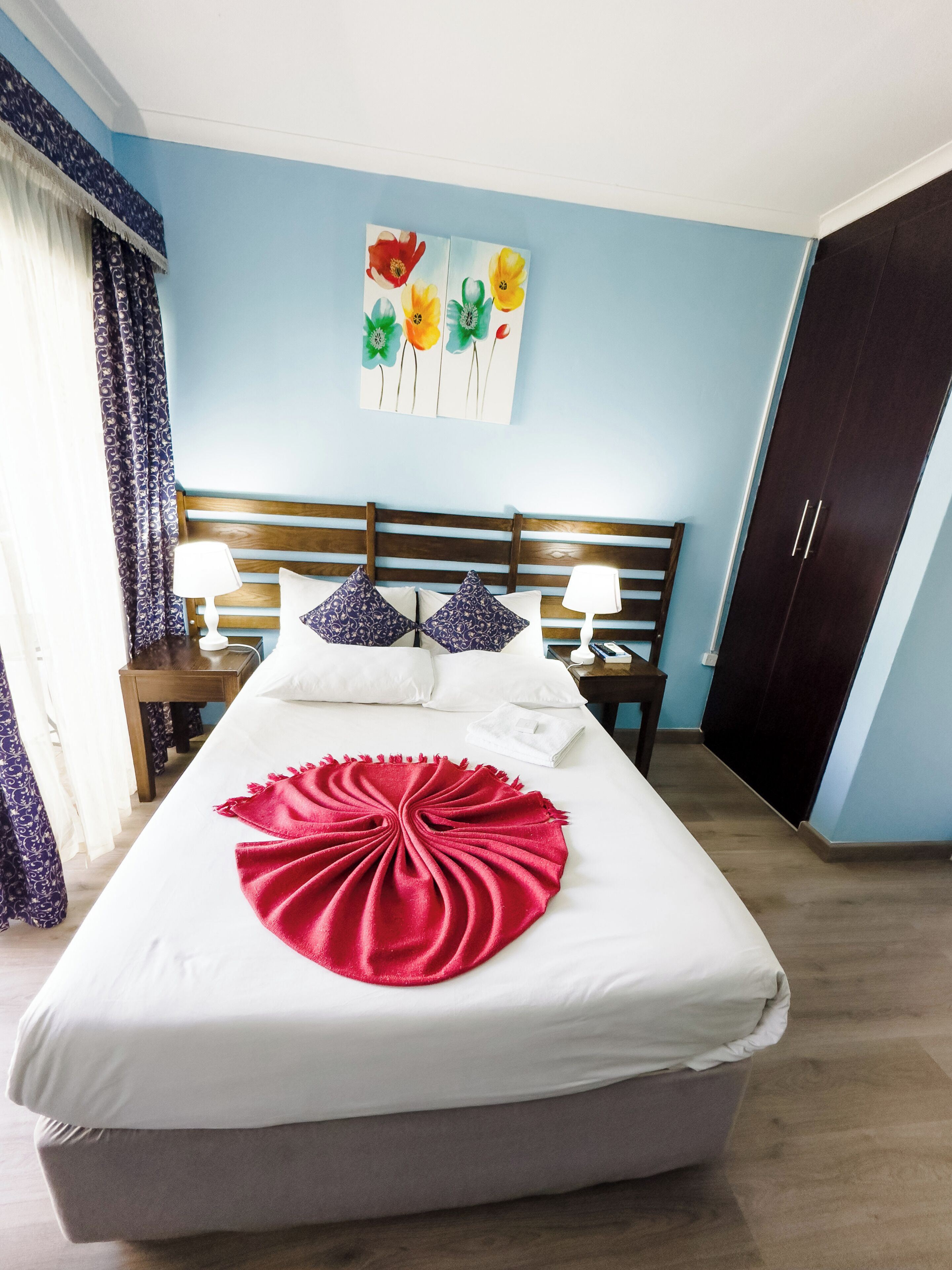 A neatly made bed with a red blanket, a wooden headboard, and a painting of flowers on the wall.