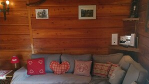 En mysig stuga med trägolv, en soffa med hjärtformade kuddar, ett rött bord och en liten träpall.
