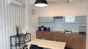 A modern kitchen with wooden cabinets, a microwave, and a kettle. There is a dining table with chairs and a black shelf with books and a plant. - Adler Bed & Breakfast (Lódz)
