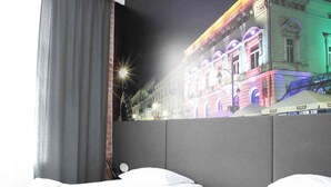 A hotel room with two beds, a large window with curtains, and a wall mural of a brightly lit building. - Adler Bed & Breakfast (Lódz)