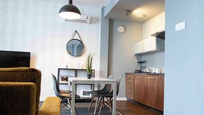 A modern living room with a sofa, a dining table with chairs, a TV, a wall-mounted mirror, and a kitchen area with cabinets and a clock. - Adler Bed & Breakfast (Lódz)