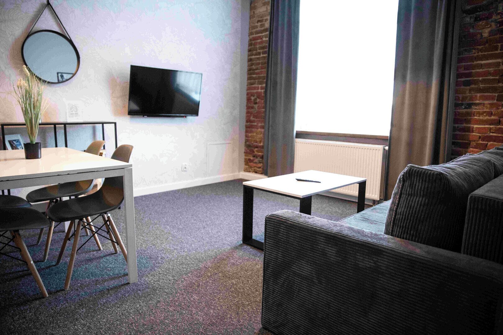 Una habitación moderna con una mesa de comedor, sillas, un sofá, una mesita de centro pequeña, un televisor de pantalla plana y un ventanal grande con cortinas.